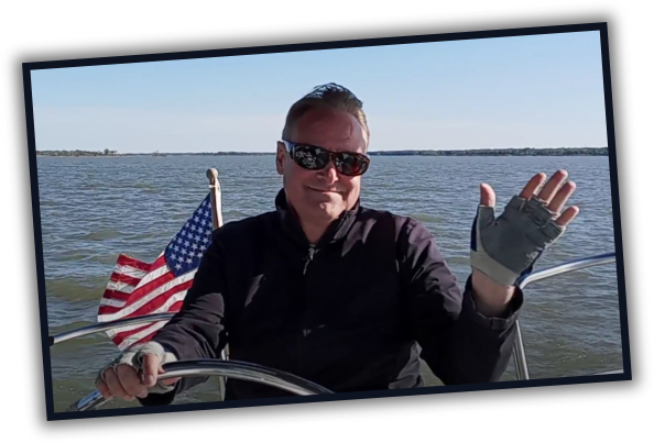Stephen at the helm of his sailboat on a sunny day, waving, with an American flag flying at the stern