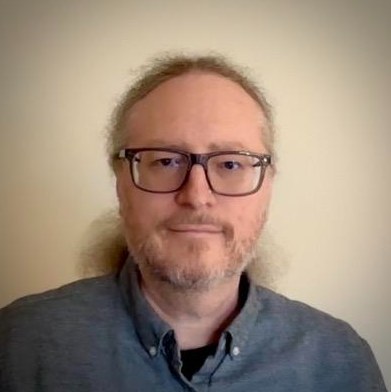 Portrait of poet David Hershhorn, smiling slightly, with glasses, a beard and long hair pulled back, wearing a gray button-down shirt
