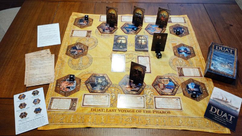 The Duat game laid out on a table: the golden Egyptian-themed game mat with ship location tiles, passenger standees, Ren and Ma’at card decks, scenario cards, dice, the game box and the quick-start booklet